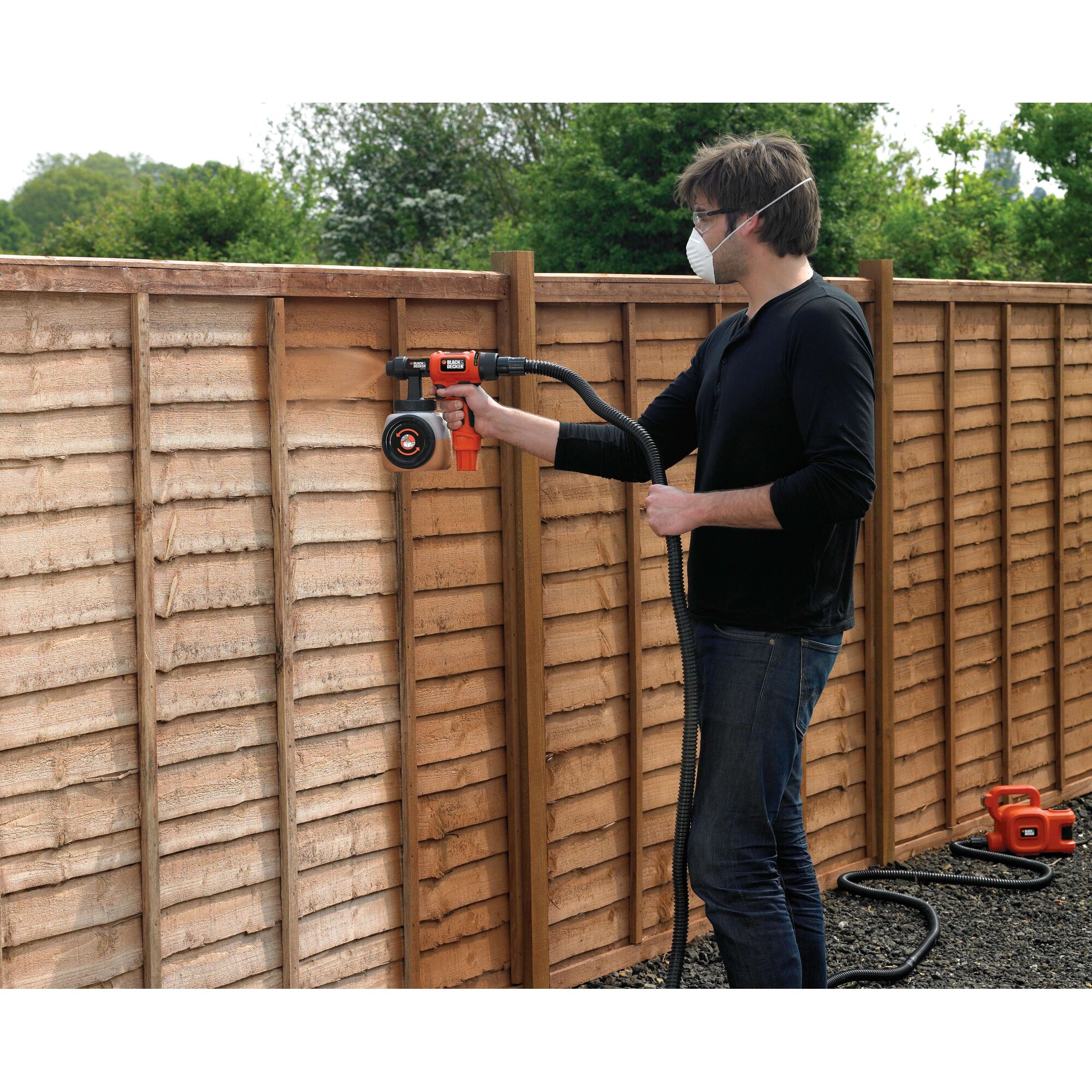 A person using a BLACK+DECKER HVLP400 paint sprayer to paint a wooden fence outdoors. The sprayer is connected to a hose and a base unit placed on the ground.