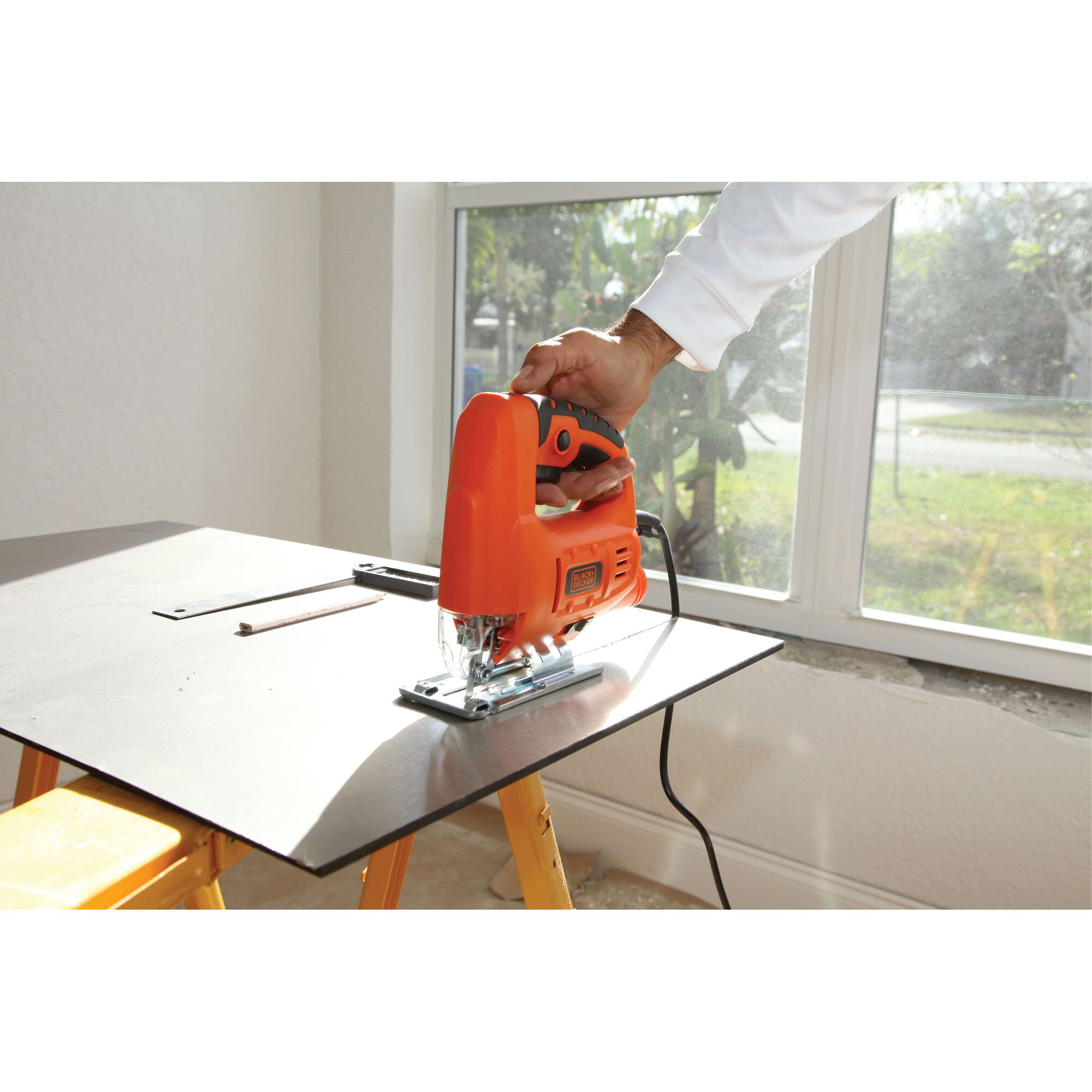 Photo of BLACK+DECKER JS20 jigsaw in use, orange body, cutting wood sheet indoors.