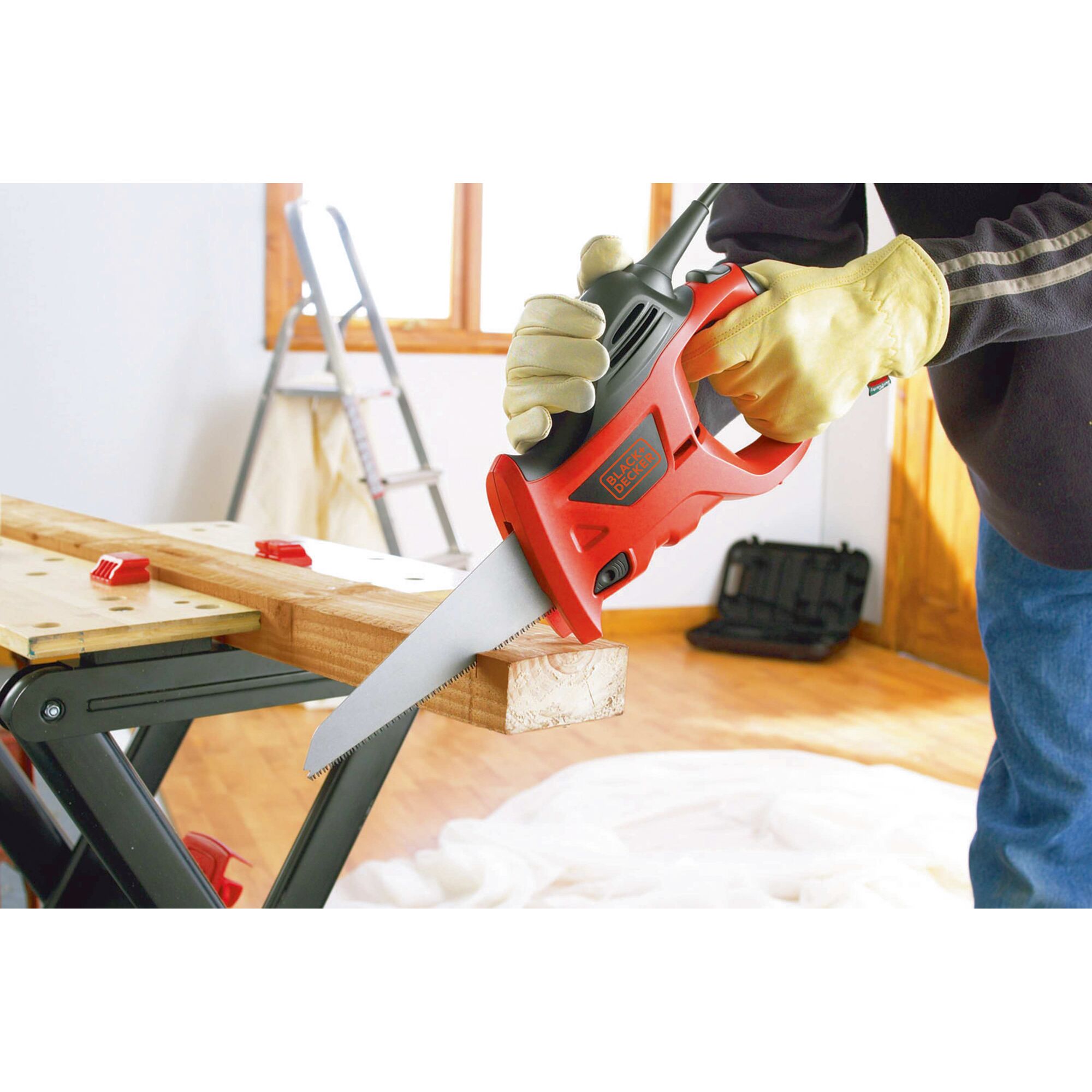 A person wearing yellow work gloves is using a BLACK+DECKER reciprocating saw to cut a piece of wood placed on a workbench. A ladder and a toolbox are visible in the background.