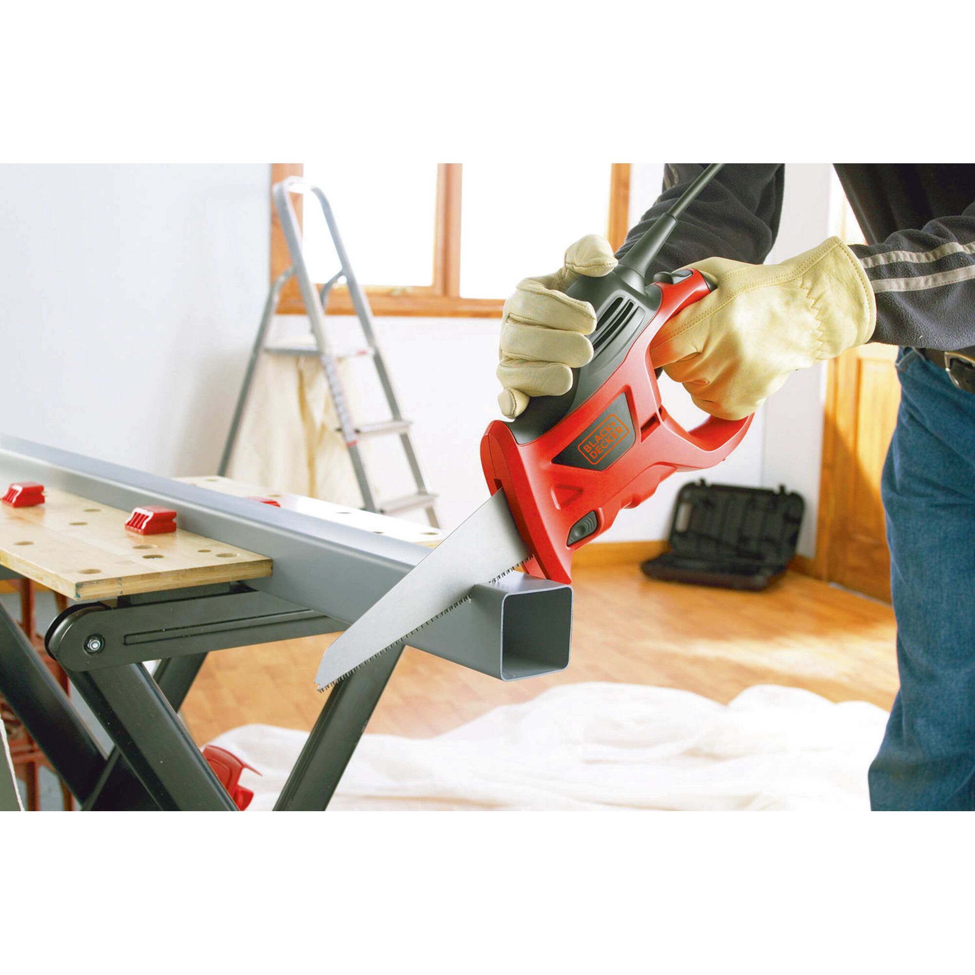 A person wearing gloves is using a BLACK+DECKER electric saw to cut a metal pipe, with a workbench and ladder visible in the background.
