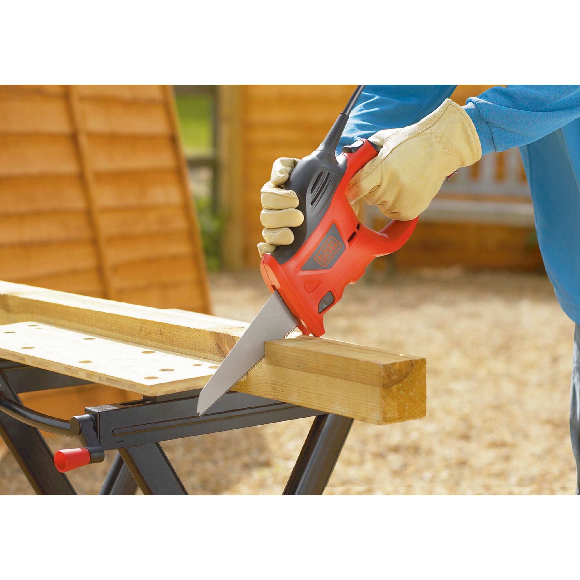 A person wearing protective gloves is using a BLACK+DECKER electric saw to cut a wooden plank on a workbench outdoors.