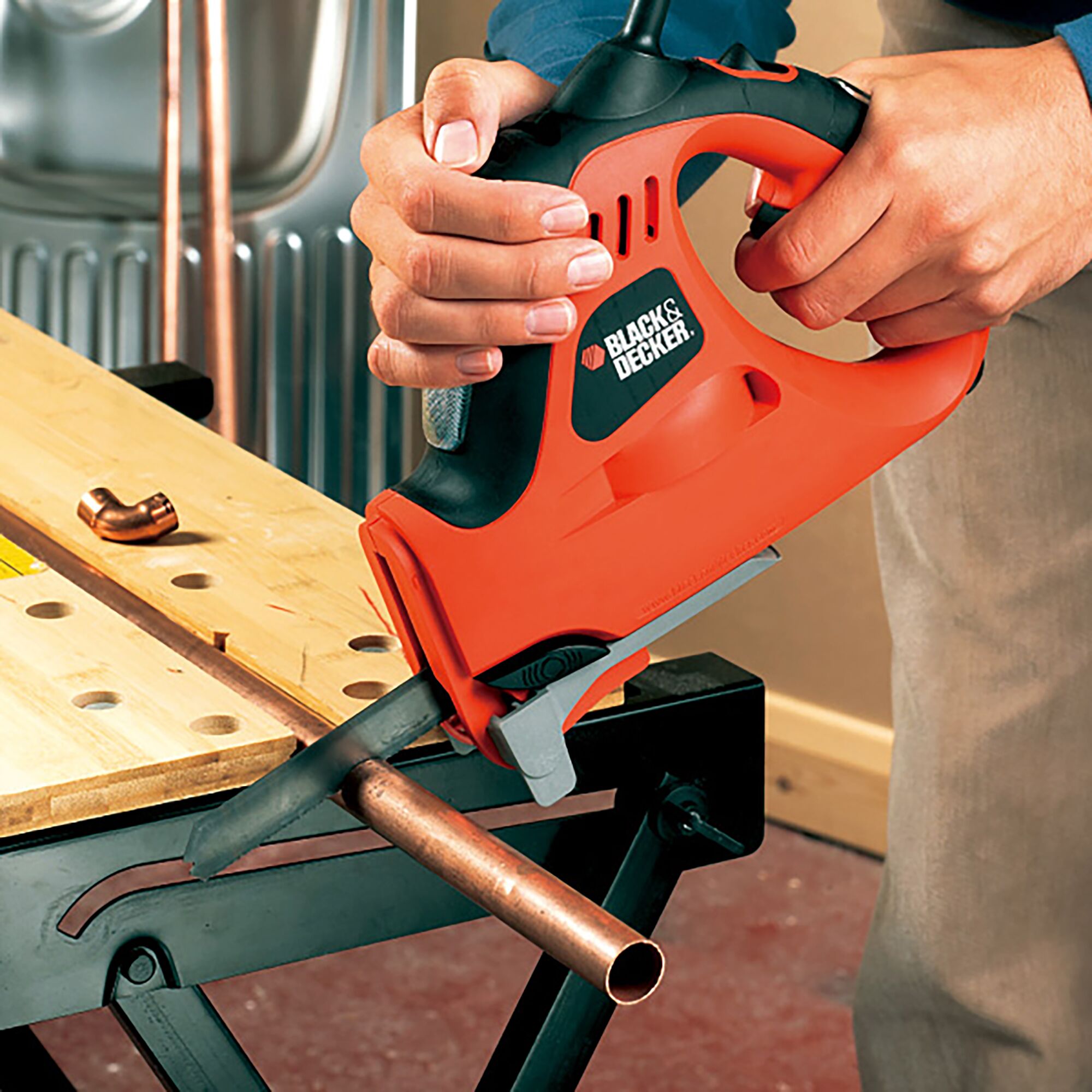 BLACK+DECKER KS900G orange and black jigsaw cutting copper pipe on workbench, close-up photo.