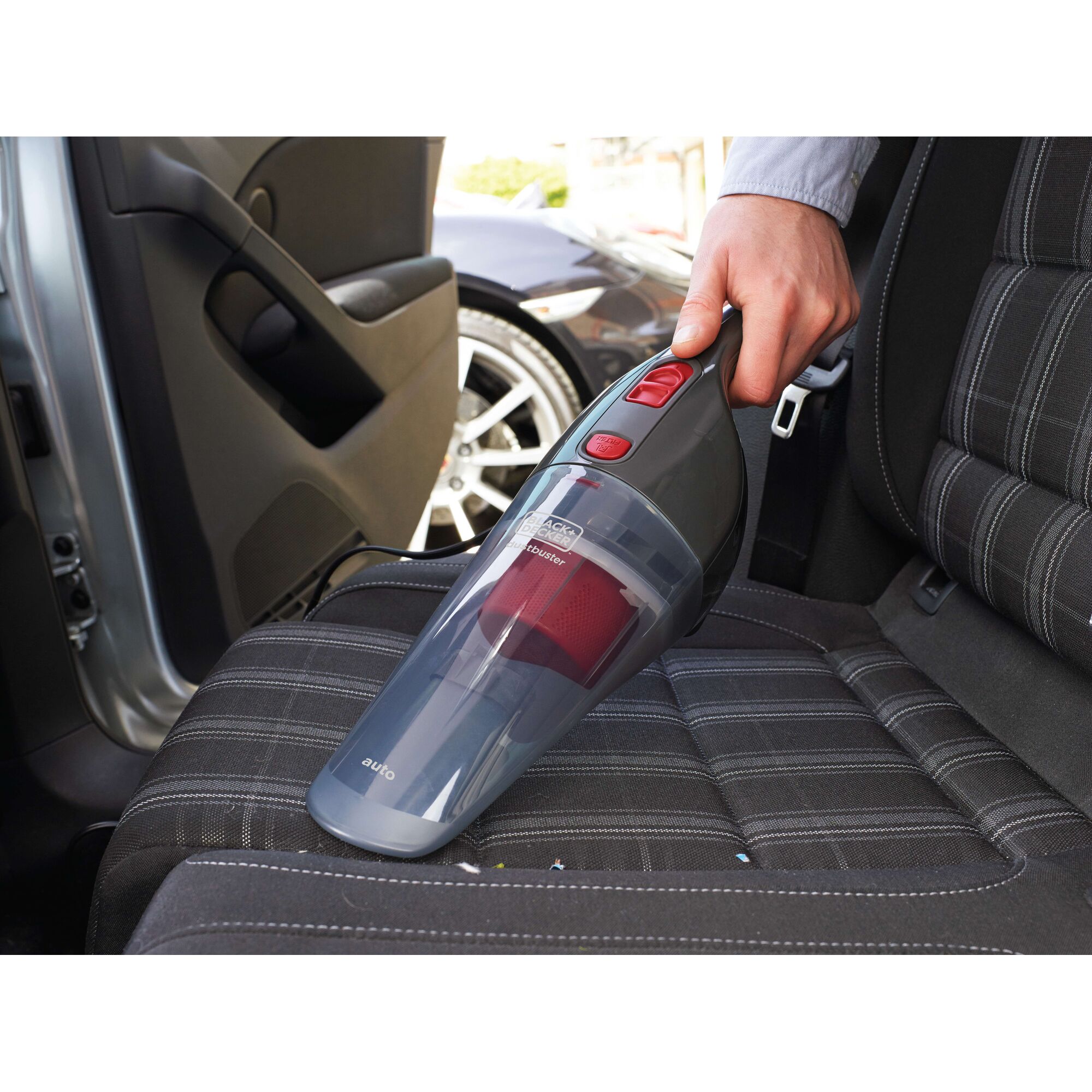 A handheld BLACK+DECKER vacuum cleaner is being used to clean the seat of a car. The device is grey with red buttons, and is shown removing dirt from the fabric seat.