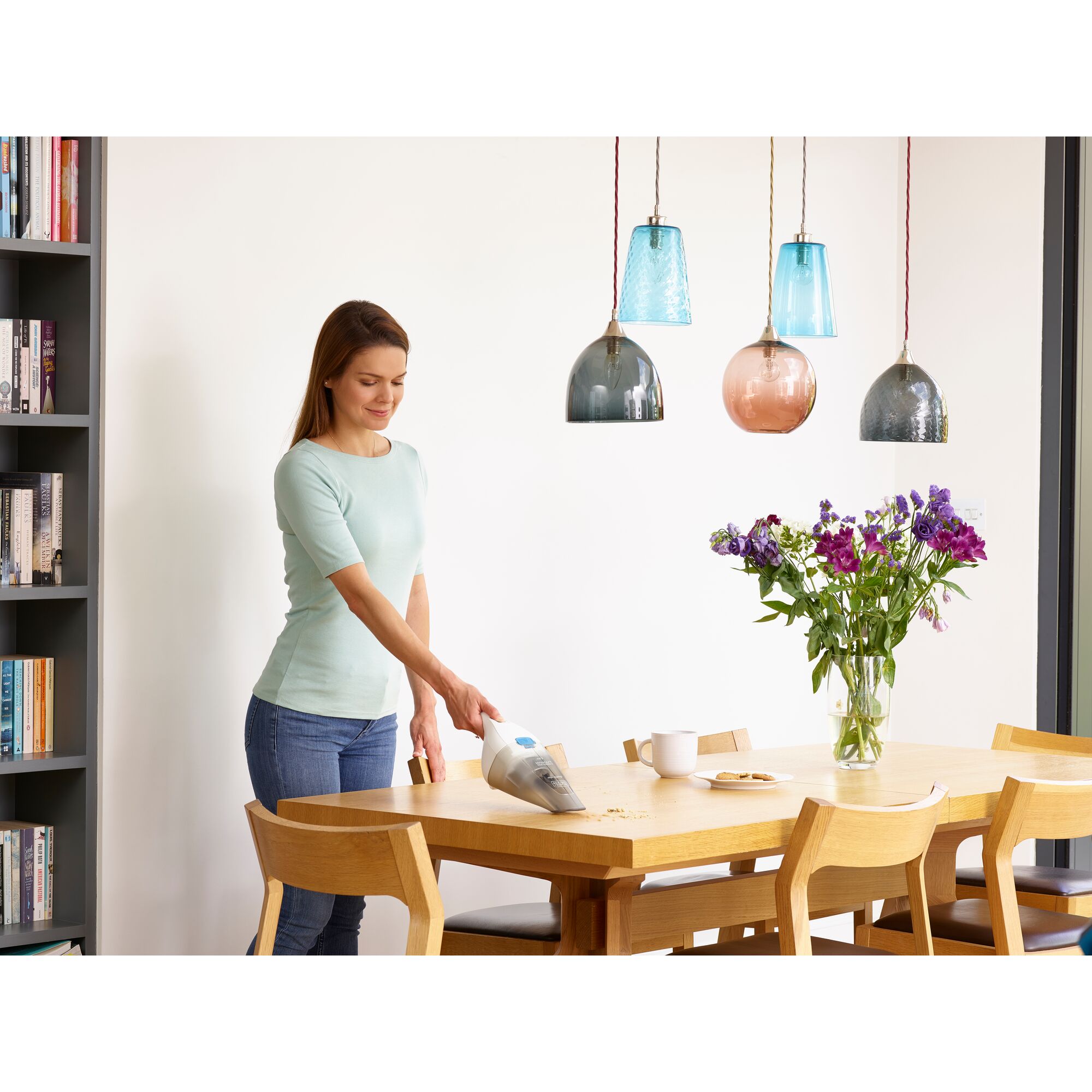 A person using a BLACK+DECKER handheld vacuum cleaner to clean crumbs from a wooden dining table in a modern home setting. There are flowers, cups, and plates on the table, and pendant lights hanging overhead.