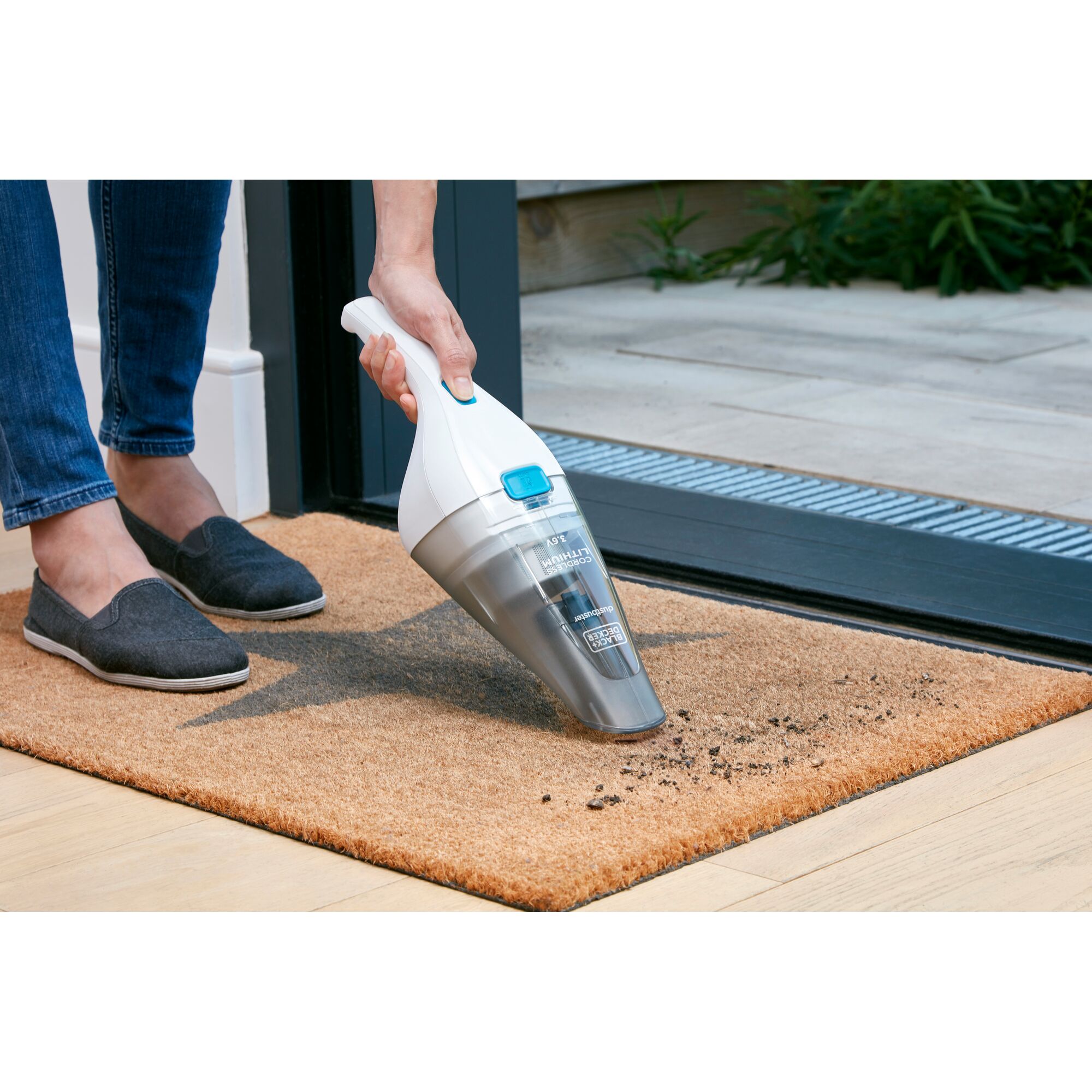 A person using a BLACK+DECKER handheld vacuum cleaner to clean dirt from a doormat placed at an open doorway.