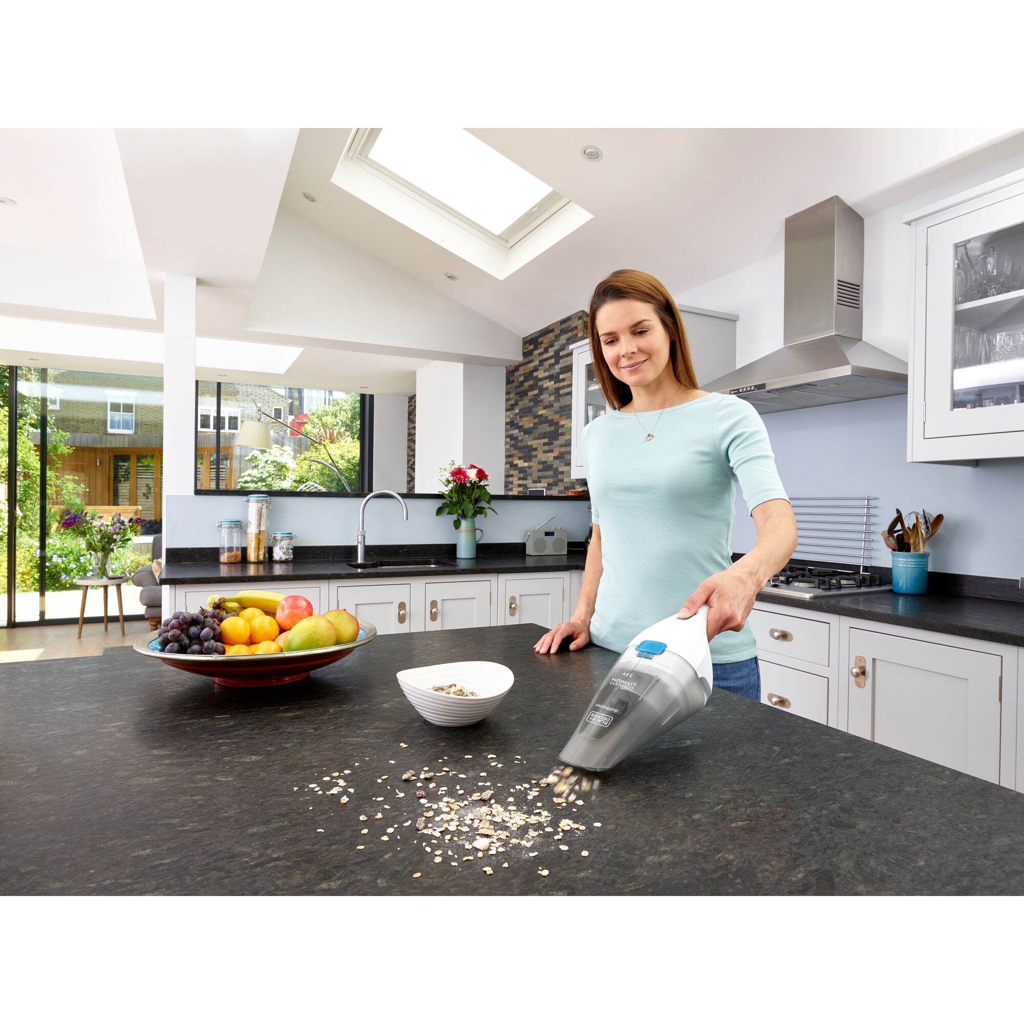 Person using a BLACK+DECKER handheld vacuum cleaner (model NVC115JL) to clean spilled oats on a black kitchen countertop, with a bowl and a fruit plate in the background.