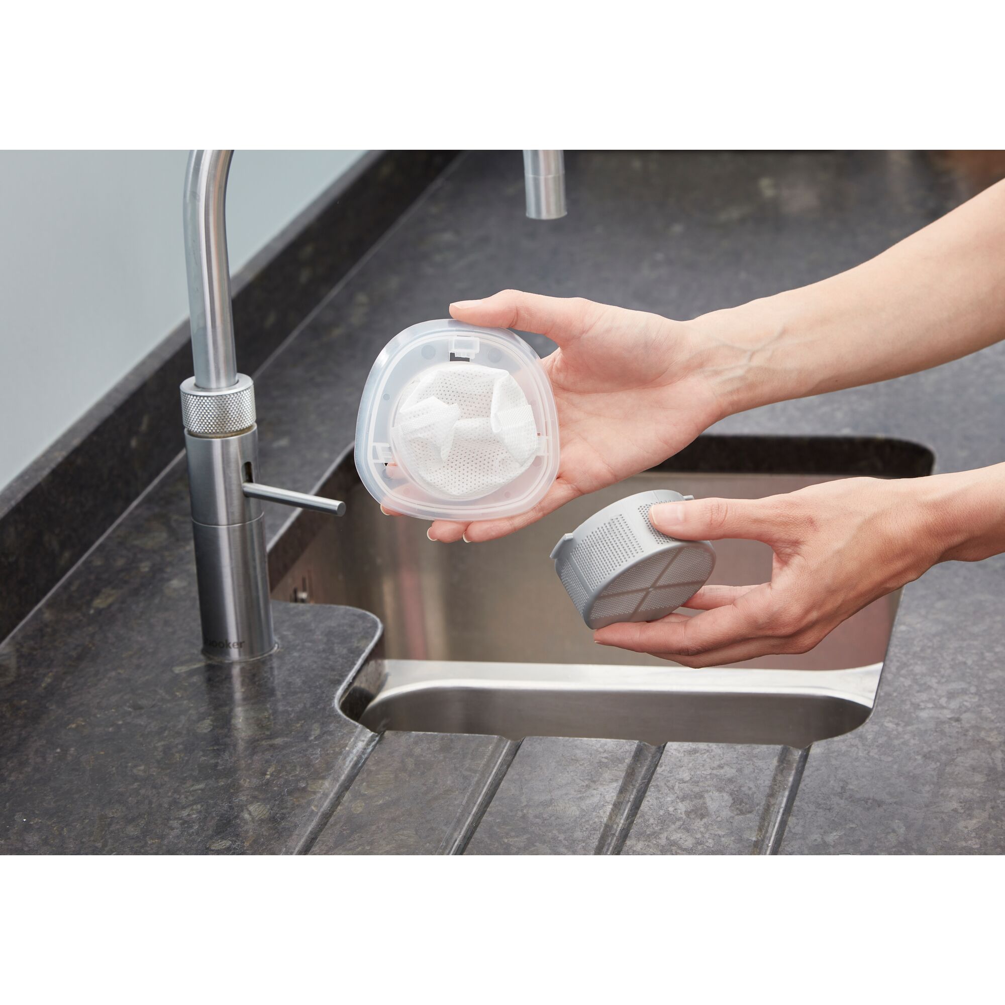 A person holds two parts of a filter from a BLACK+DECKER product above a kitchen sink. One part is transparent and contains a mesh filter.