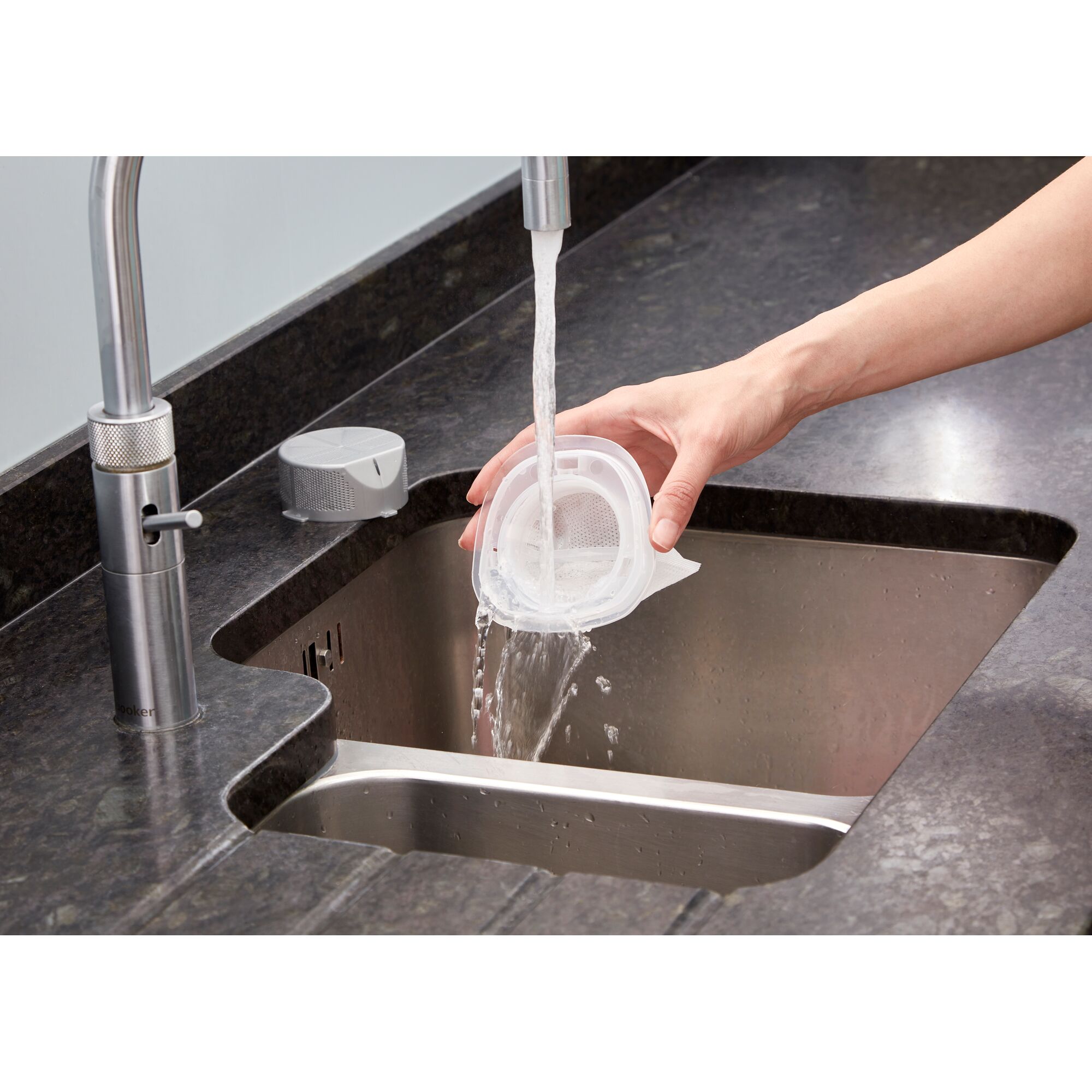 A hand rinsing a small, white plastic filter under running water at a kitchen sink, with a dark countertop and a silver faucet visible.