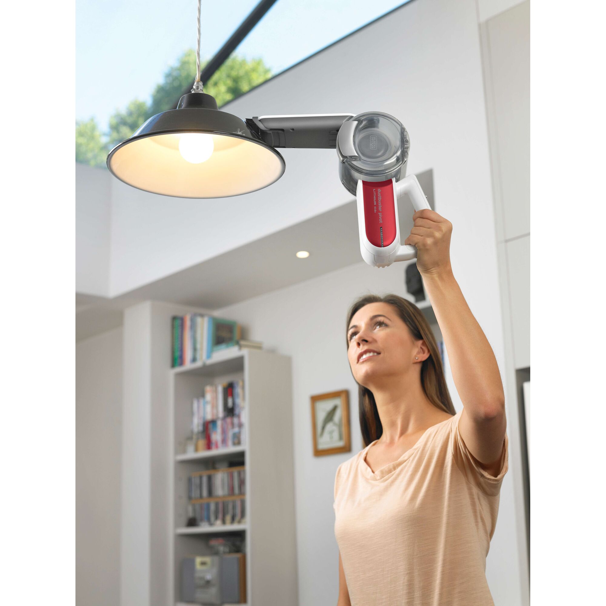 A person using a BLACK+DECKER PV1020LR handheld vacuum cleaner to clean a ceiling lamp in a modern living room. Their face is blurred for privacy.