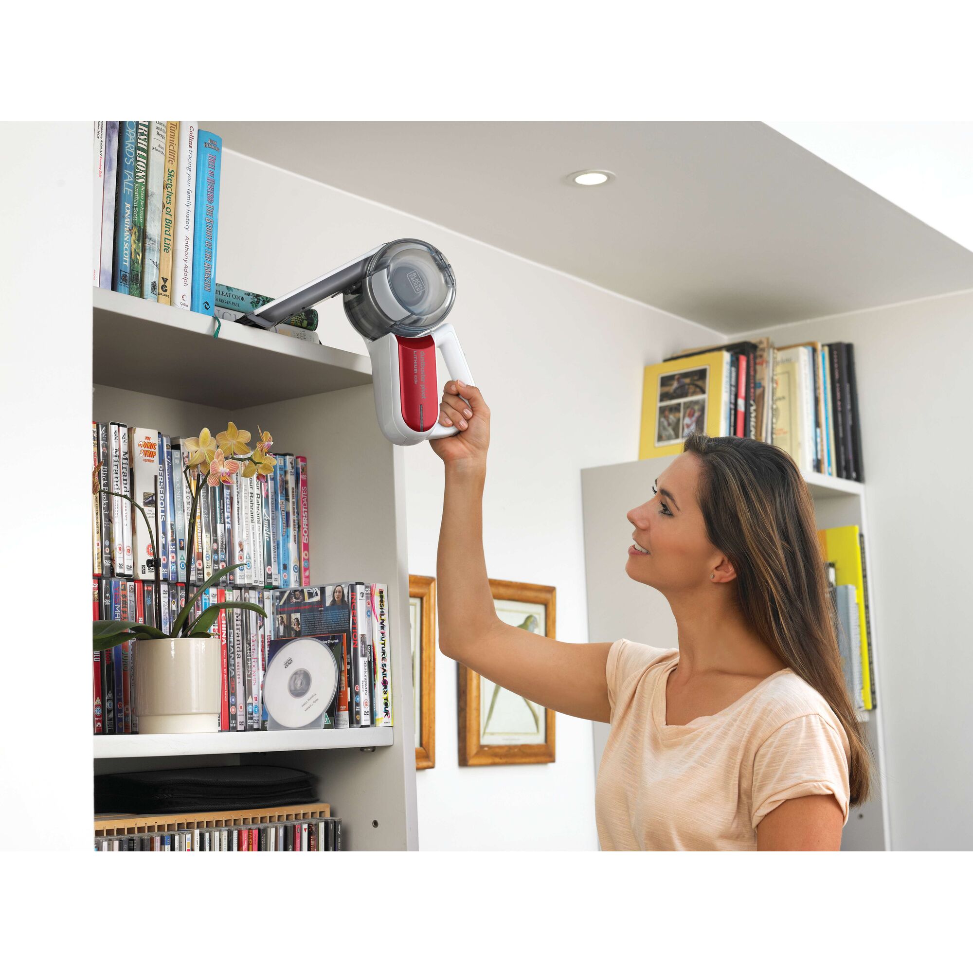A person using a BLACK+DECKER handheld vacuum cleaner to clean dust from the top shelf of a bookcase, which is filled with books and DVDs. The person's face is blurred for privacy.