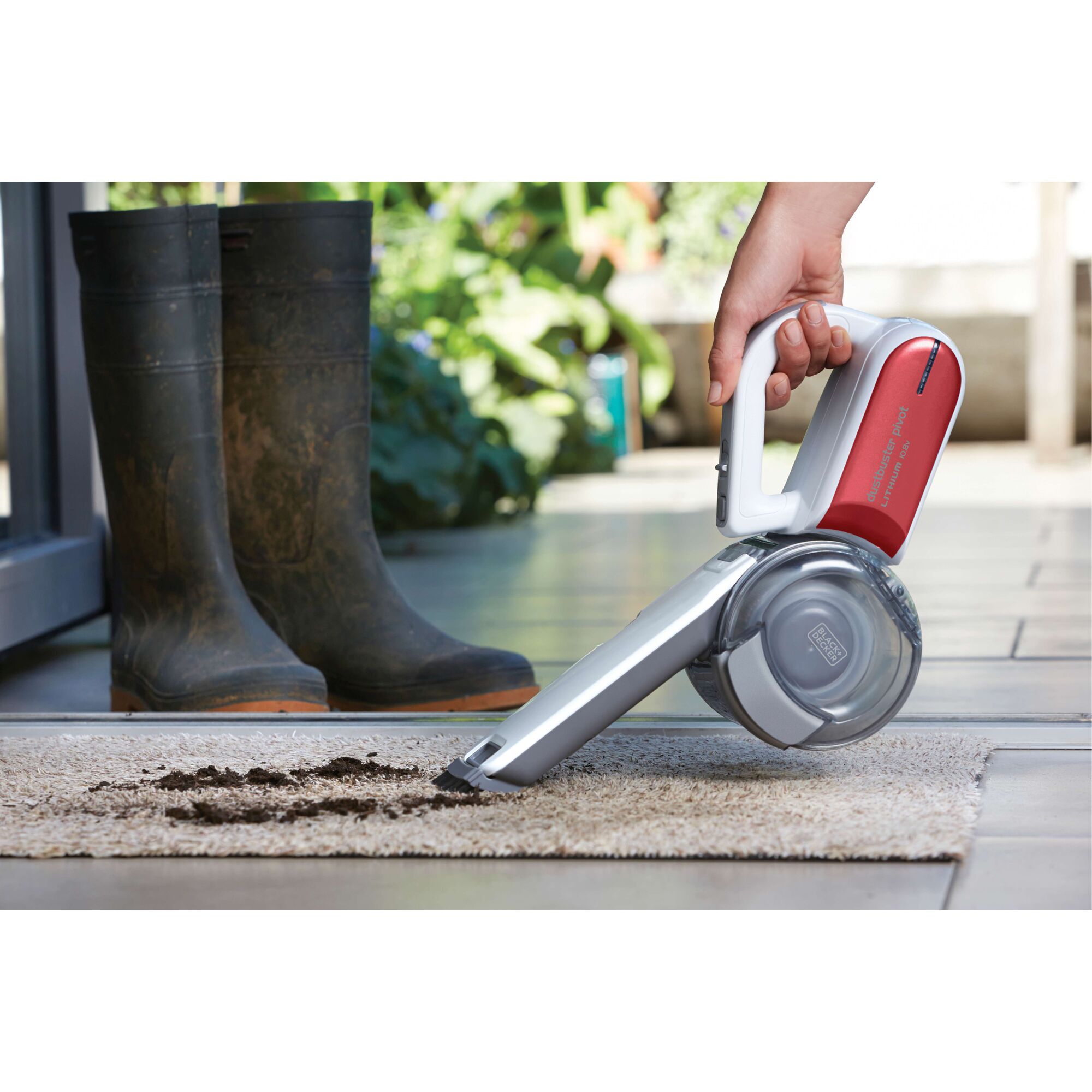A person is using a BLACK+DECKER handheld cordless vacuum cleaner (model PV1020LR) to clean dirt from a light-colored rug near a pair of rubber boots, indoors by a garden entrance.