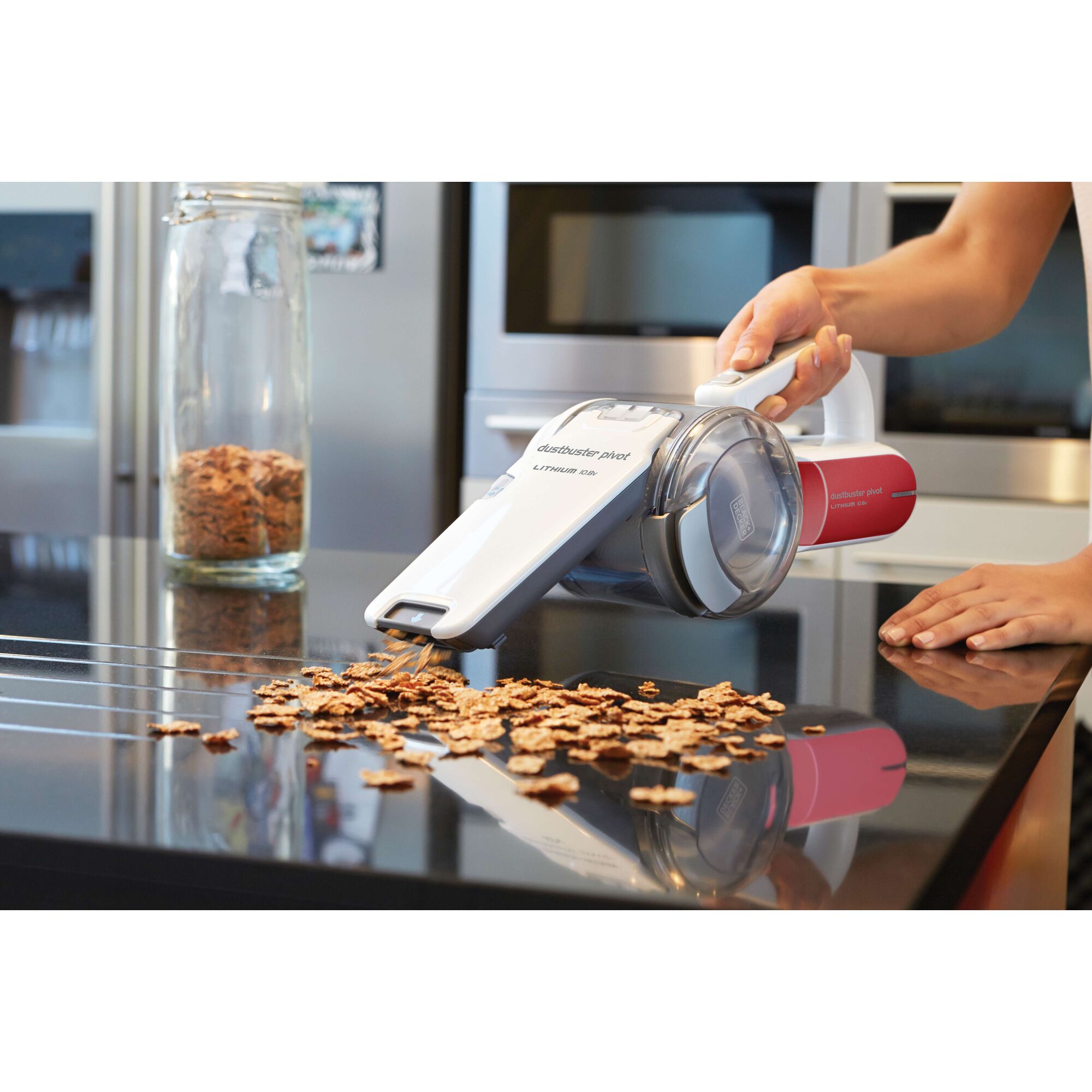 A person uses a BLACK+DECKER handheld vacuum cleaner (model PV1020LR) to clean cereal pieces from a shiny kitchen countertop. There is a glass jar containing cereal in the background.