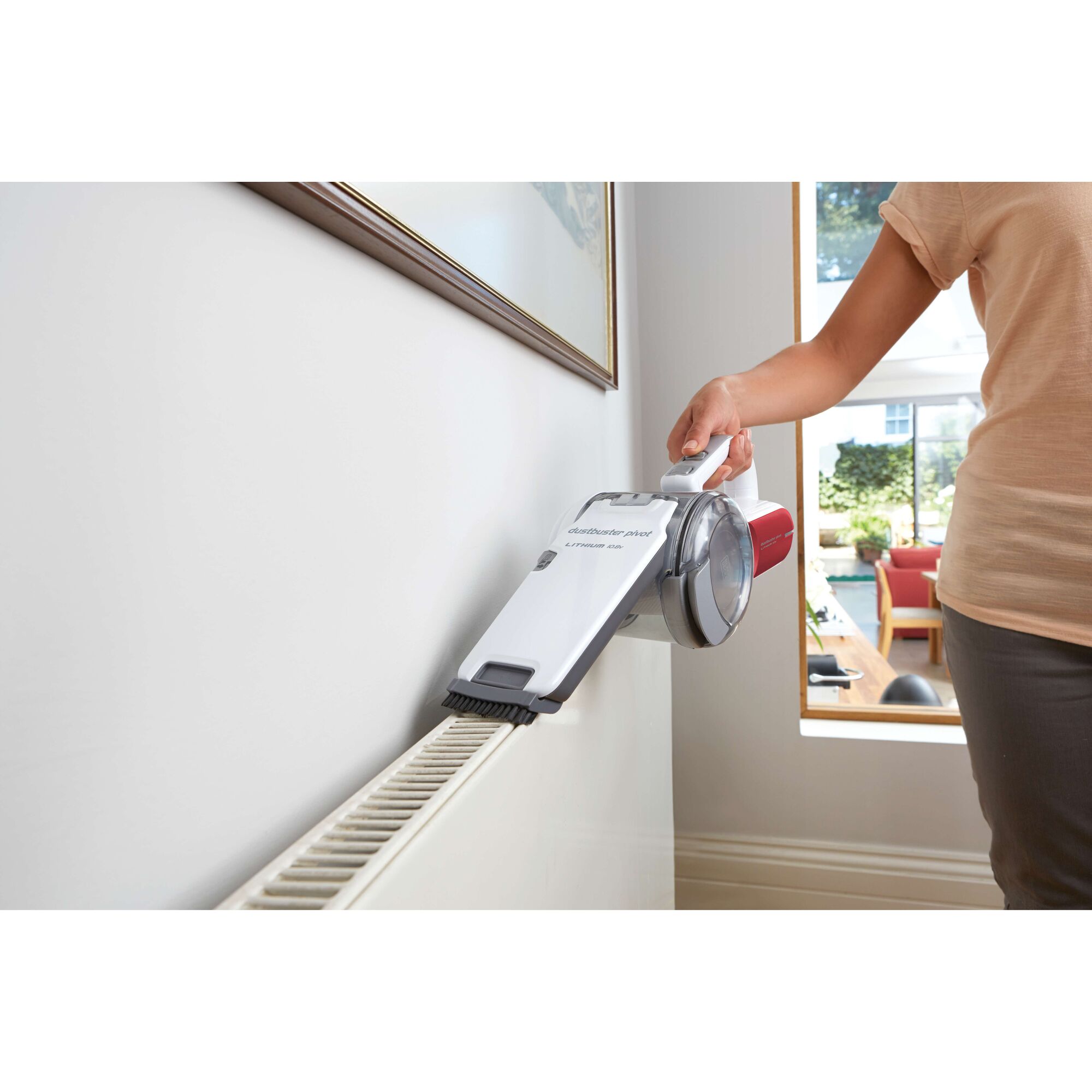 A person using a BLACK+DECKER handheld vacuum cleaner to clean a radiator in a modern home.