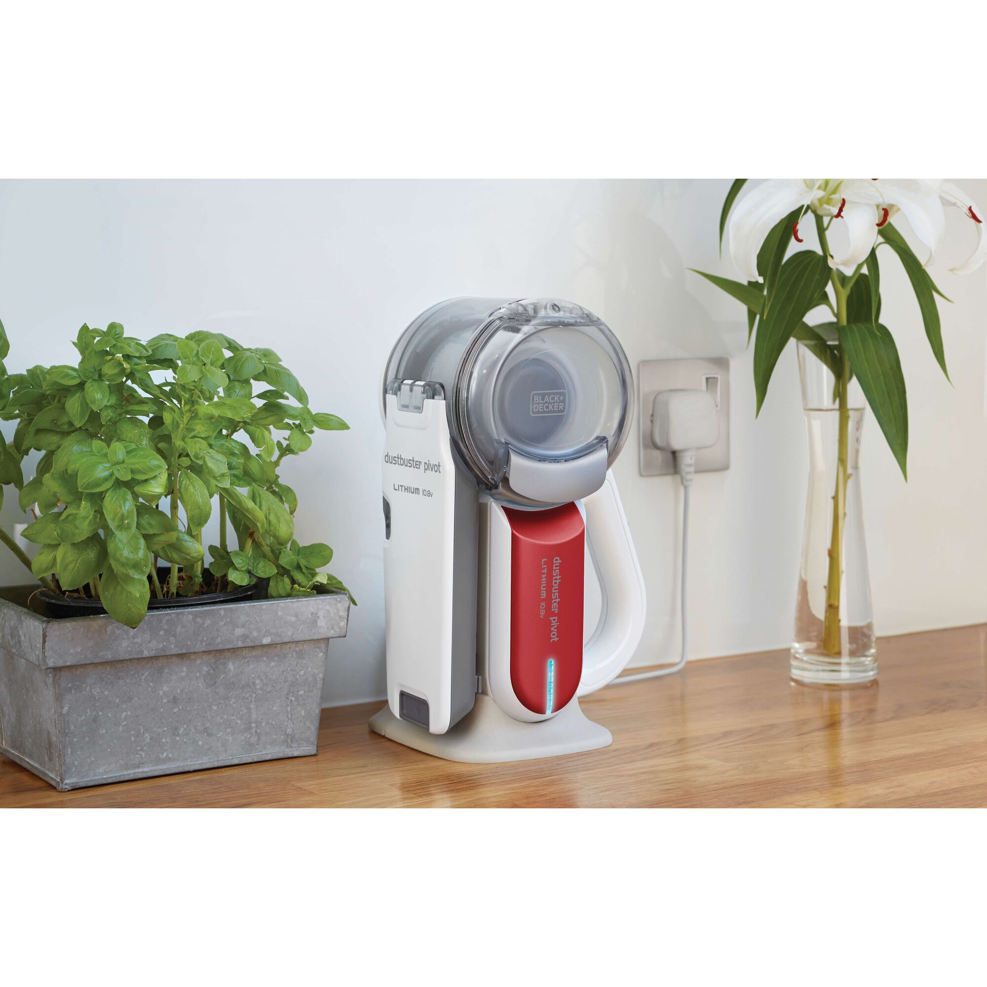 A BLACK+DECKER Dustbuster Pivot lithium handheld vacuum cleaner is shown charging on a wooden countertop. Next to it is a metal planter with green plants and a glass vase with white lilies, with a power outlet visible in the background.