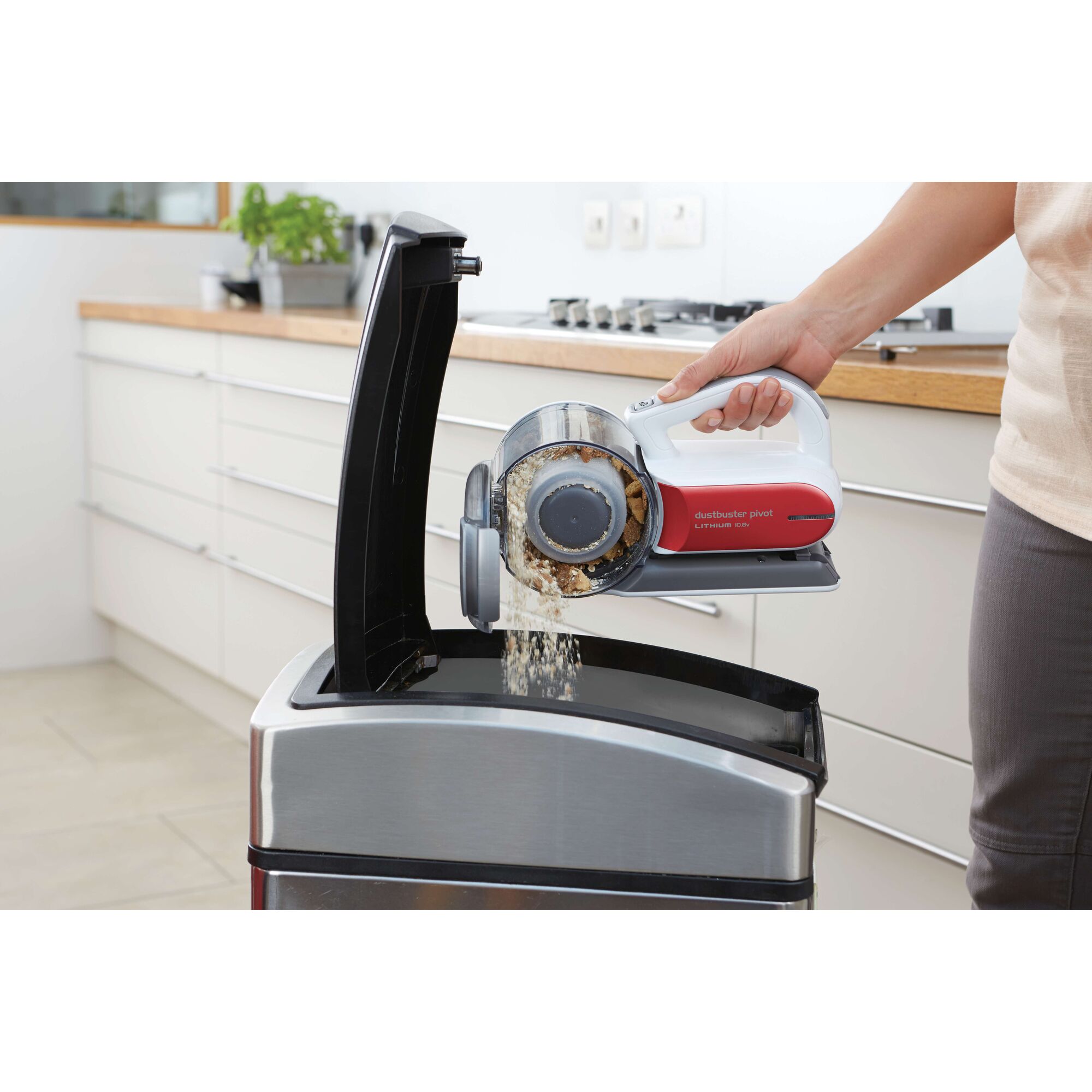 A person is emptying the dust container of a BLACK+DECKER handheld vacuum cleaner into a kitchen trash bin.