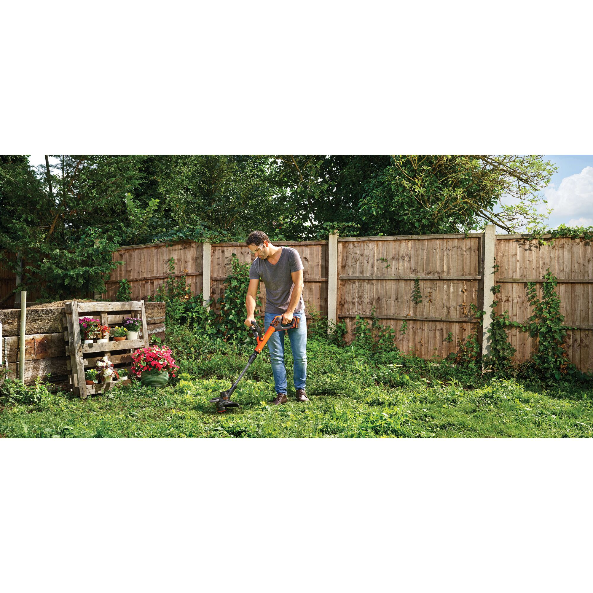 BLACKDECKER STC1840EPC cordless grass trimmer in use in a garden setting with wooden fence and plants.