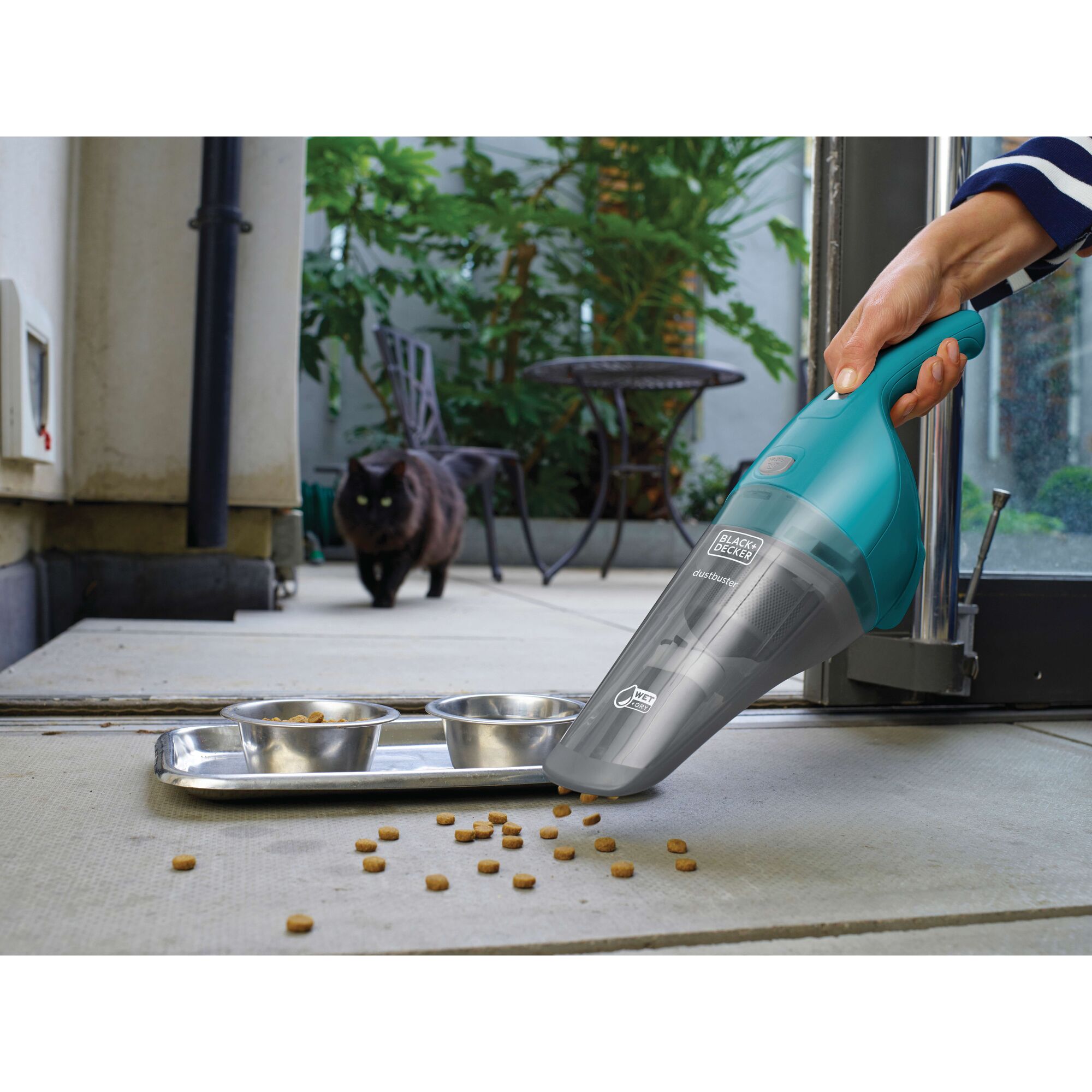 A person uses a BLACK+DECKER handheld vacuum cleaner to pick up pet food scattered on the floor near a metallic pet food tray with bowls. In the background, a black cat is walking towards the tray in an outdoor space with plants and furniture.