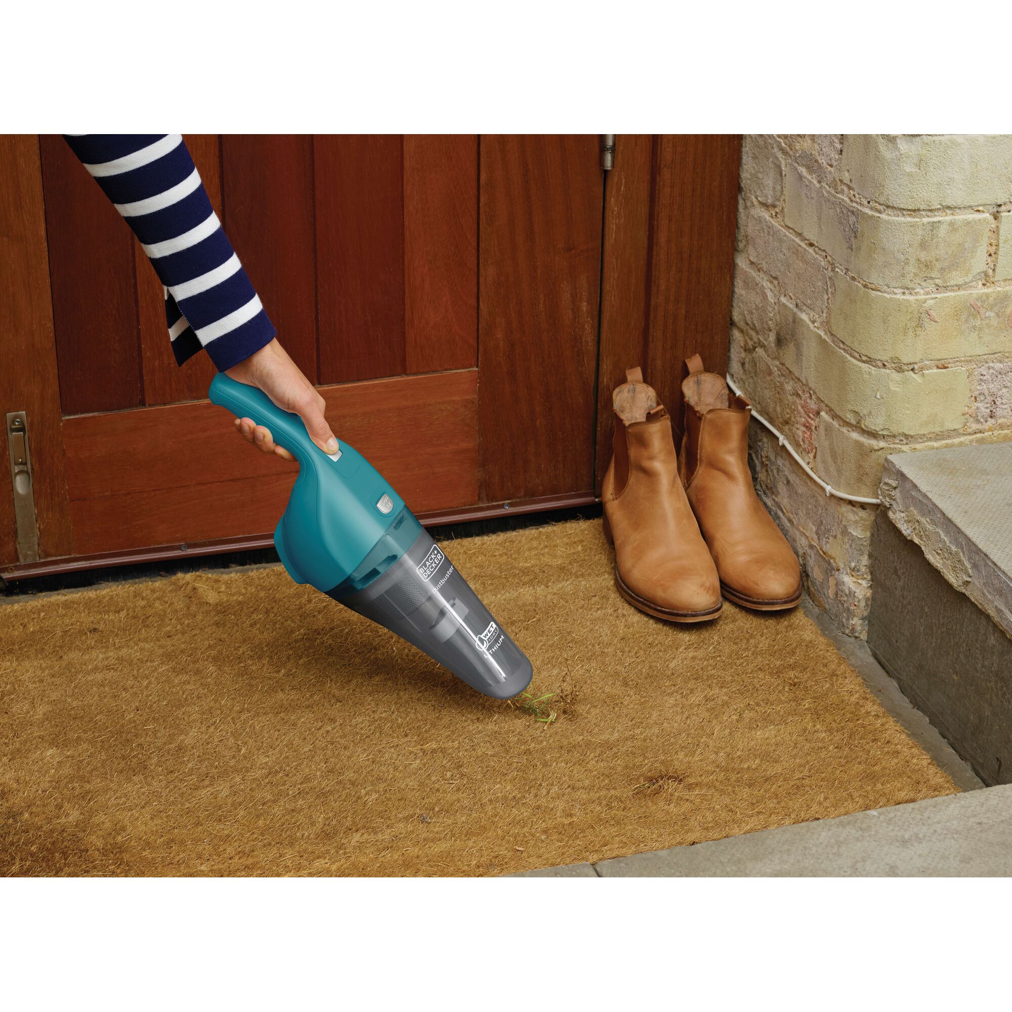 A person using a teal BLACK+DECKER handheld vacuum cleaner to clean a doormat near a wooden door, with a pair of tan boots placed beside the mat.