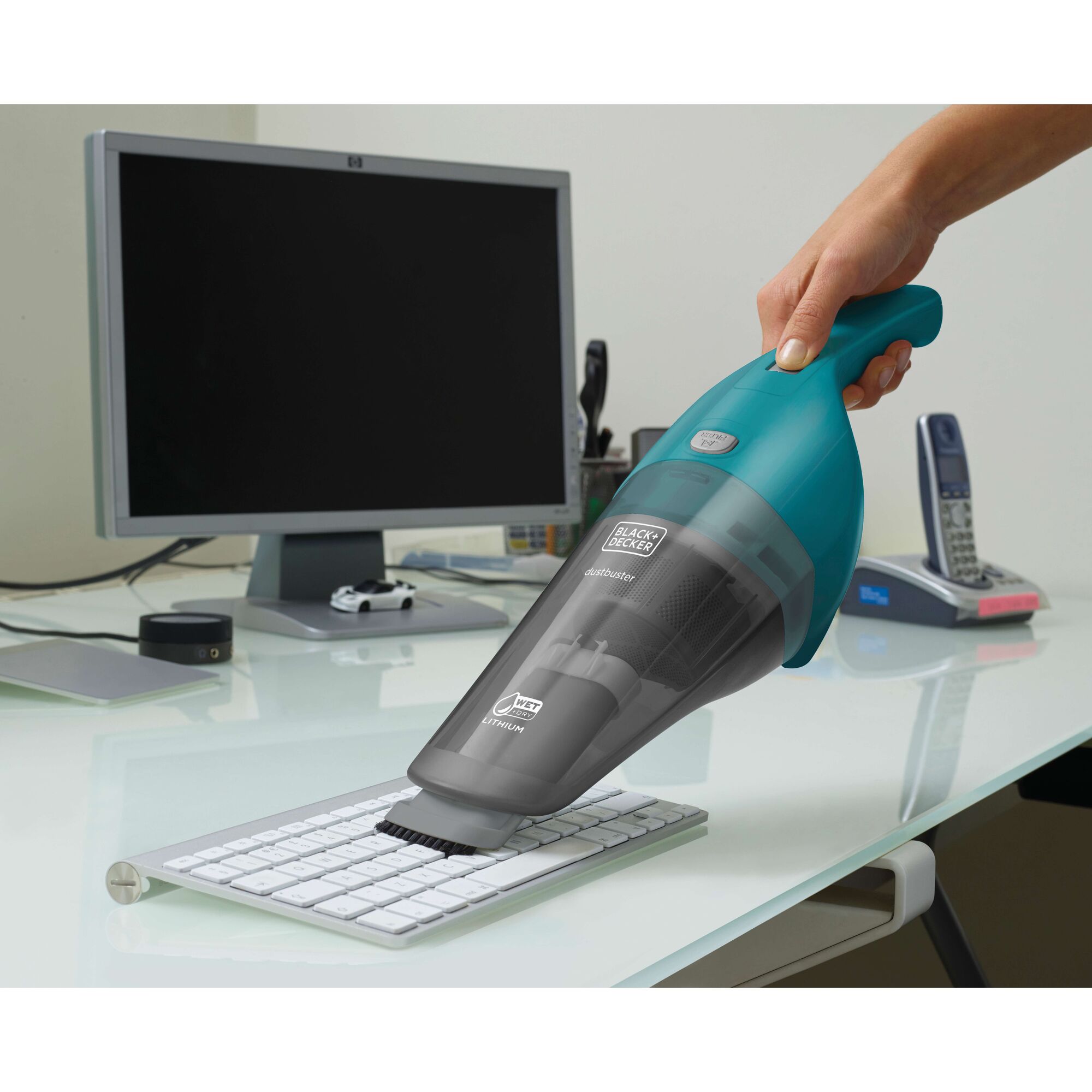 A hand is using a BLACK+DECKER handheld vacuum cleaner (model WDB115WA) to clean a computer keyboard on a desk with a computer monitor, phone, and office supplies in the background.