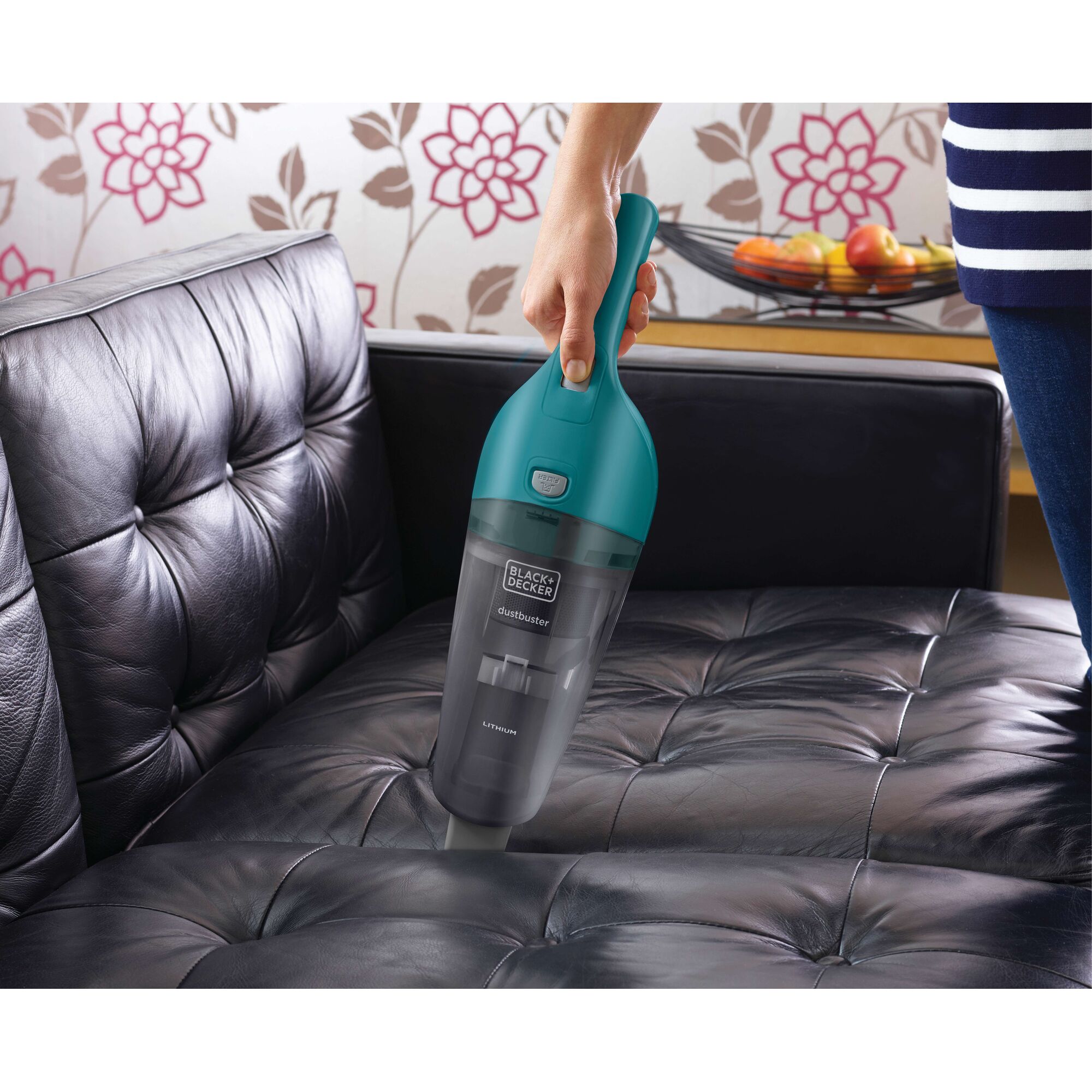 A person is using a BLACK+DECKER Dustbuster handheld vacuum cleaner to clean a black leather sofa.
