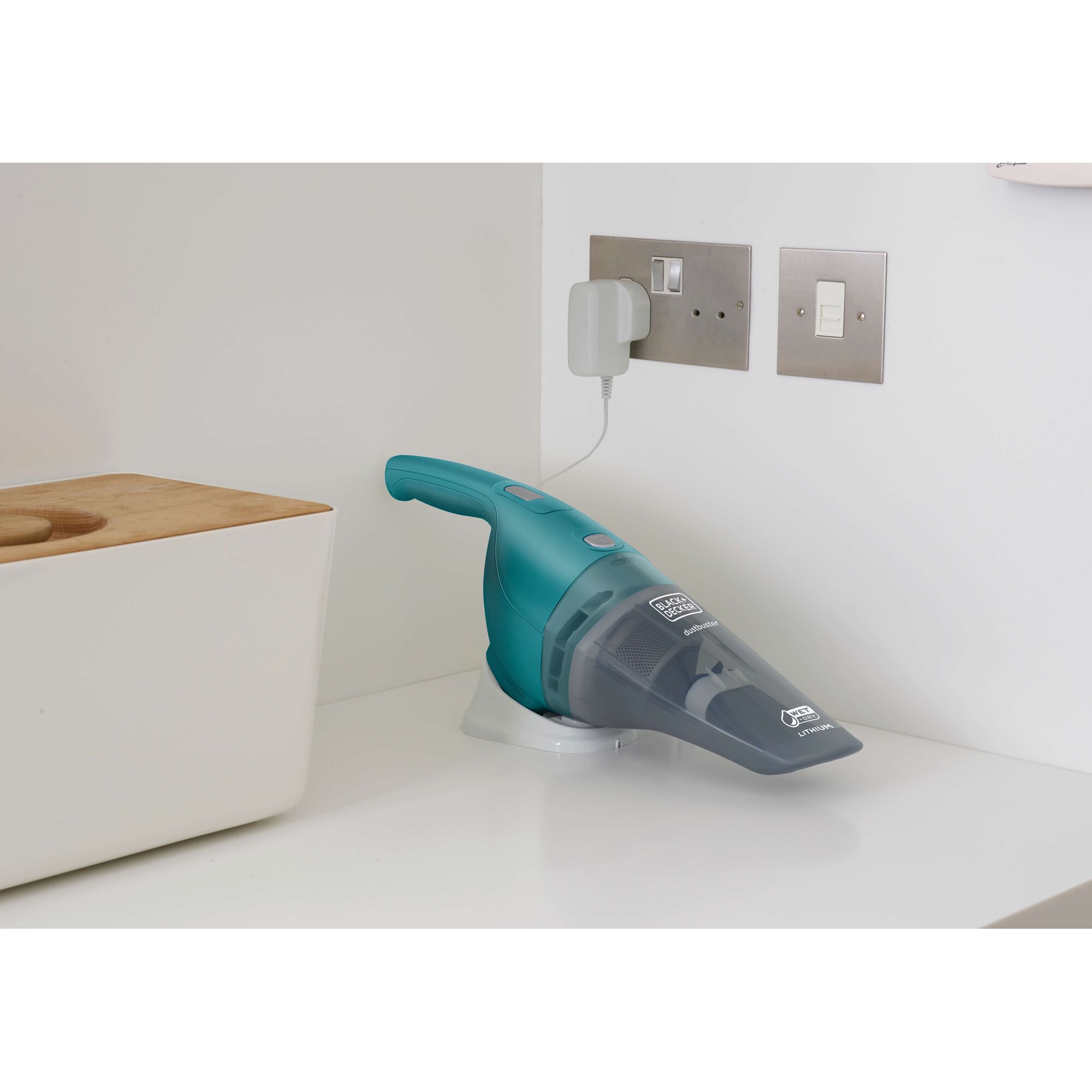 A small handheld BLACK+DECKER vacuum cleaner is placed on a white countertop, charging via a wall socket. The vacuum is turquoise and gray, and sits next to a white container with a wooden lid.
