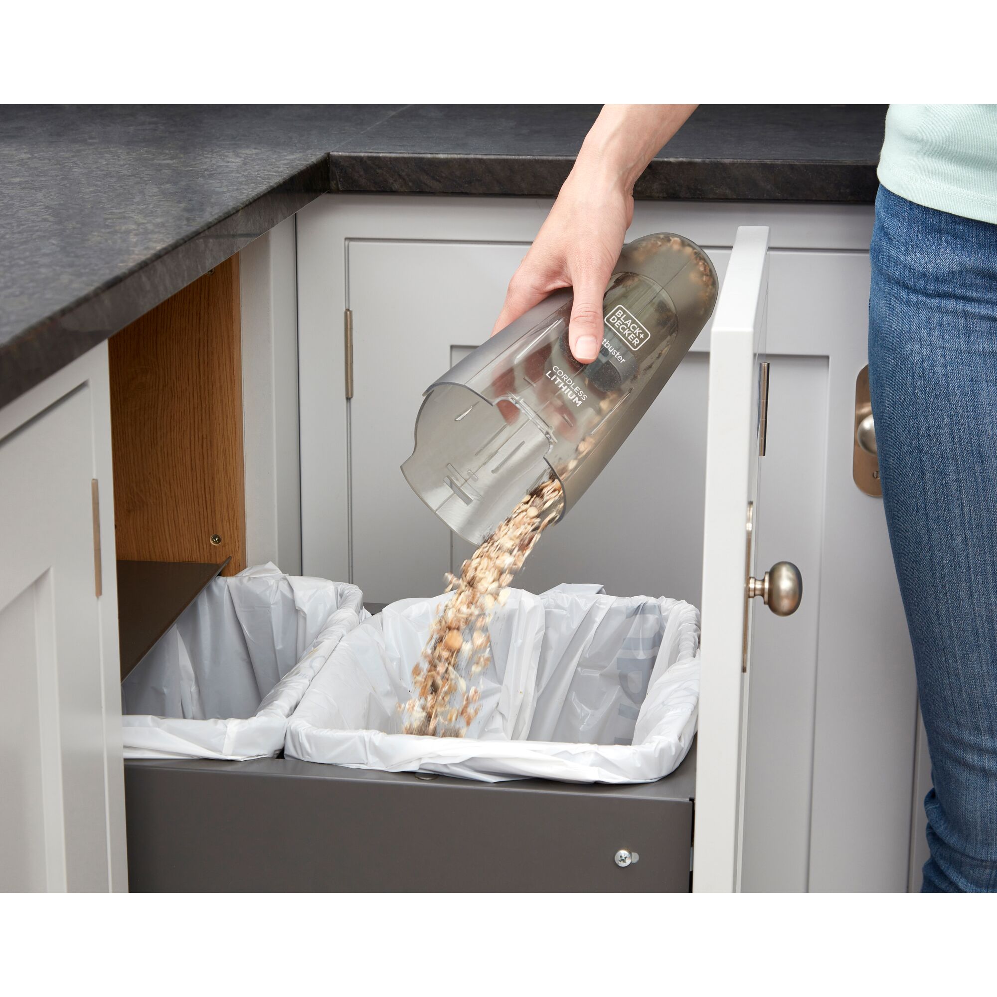 Hand emptying BLACK+DECKER WDC115W handheld vacuum canister into trash bin in kitchen.